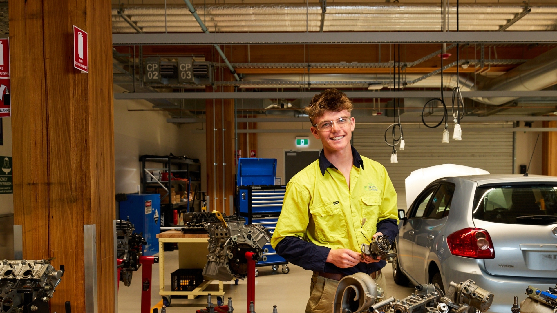 Automotive student holding a mechanical component in a workshop beside a car and engine equipme