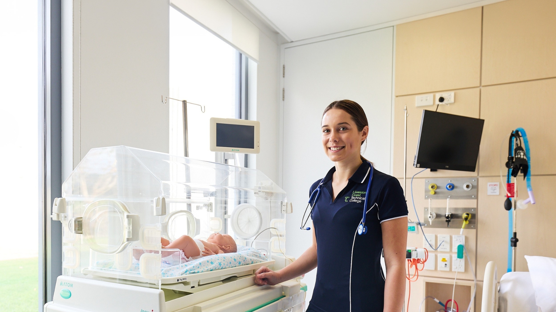 Health student standing beside a neonatal incubator in a clinical training environment.
